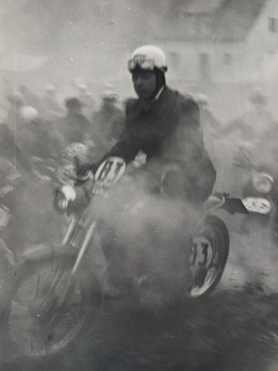 Historischer Motorradfahrer mit Helm und Brille auf einer Rennmaschine, umgeben von Rauch